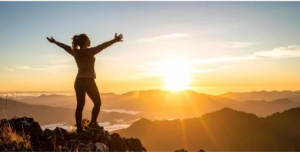 Woman greeting the sunrise with arms outstretched, atop a mountain.