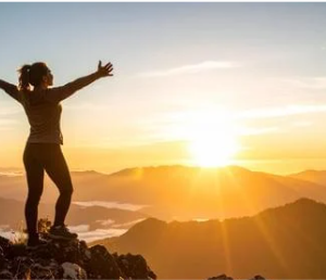 Woman greeting the sunrise with arms outstretched, atop a mountain.